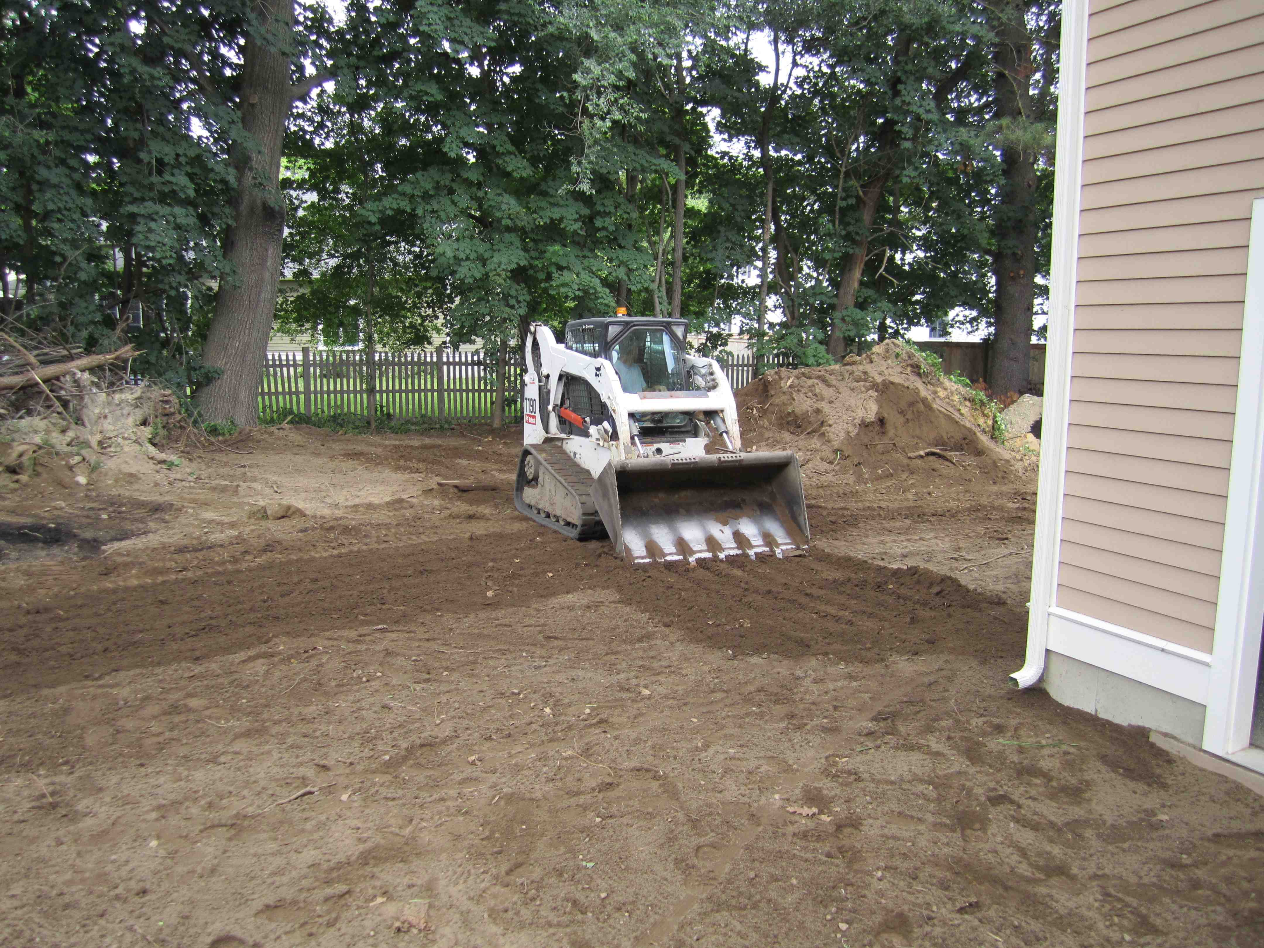 A landscaping crew uses heavy machinery to level and grade the soil in a residential backyard, preparing the ground for new lawn installation. - lawn layers near me